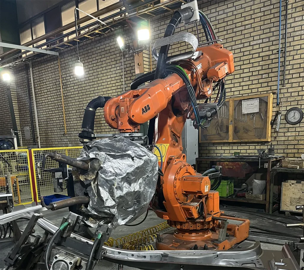 نمای نزدیک از یک ربات صنعتی ایبیبی (ABB) نارنجی رنگ کارکرده در حال کار روی یک فیکسچر با قطعه فلزی در محیط کارخانه و سوله صنعتی با دیوار آجری